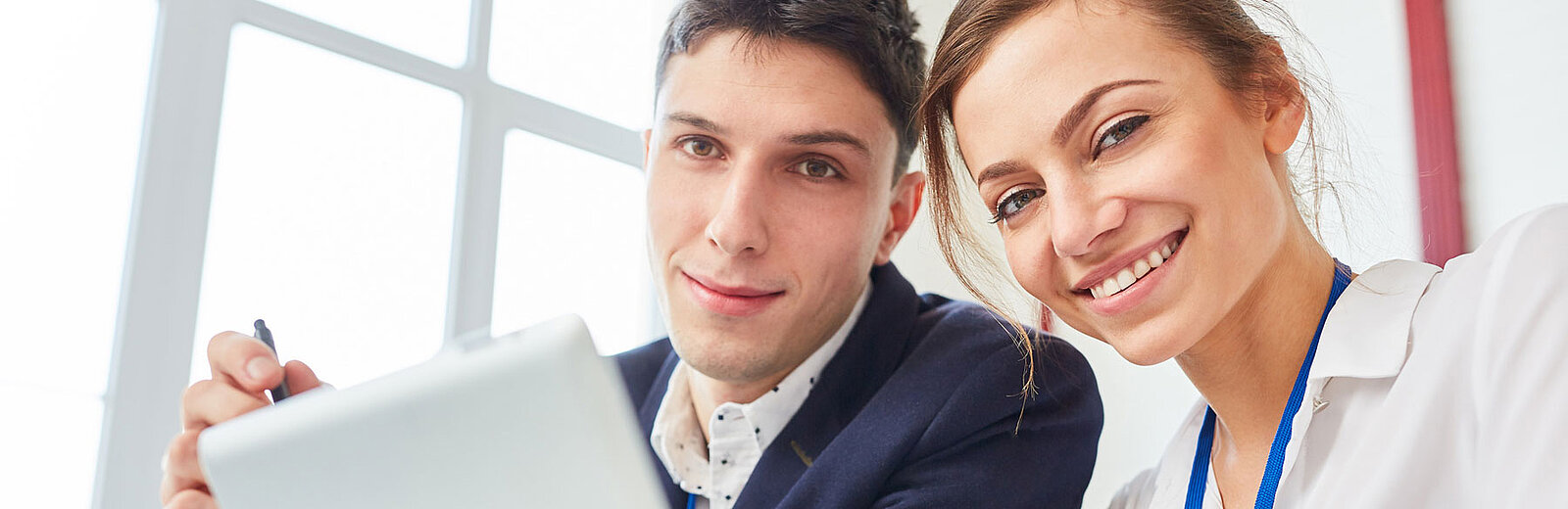 Zwei lächelnde Menschen, ein Mann und eine Frau, sitzen zusammen in einem Büro und schauen in die Kamera